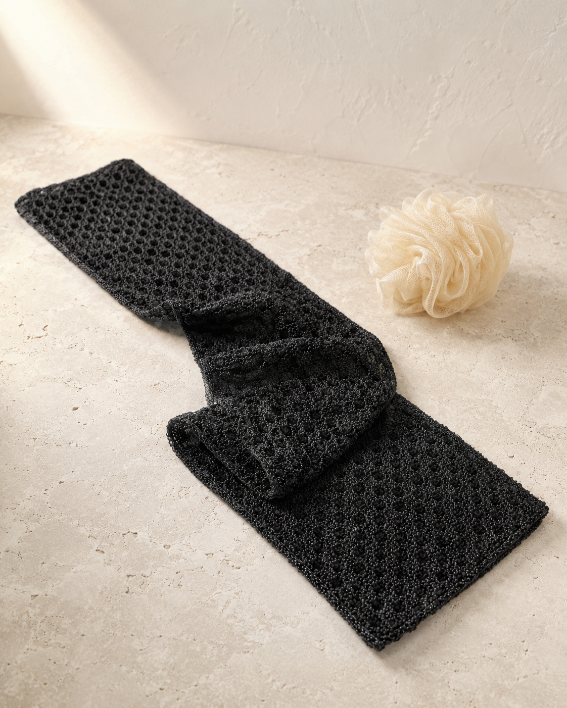 Full-length black Kuyona exfoliating towel on warm stone beside a conventional shower loofah